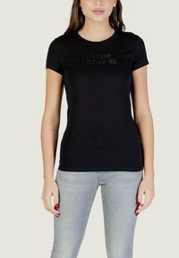 Femme portant un t-shirt noir ajusté avec un détail de texte subtil et un jean bleu clair, debout devant un fond uni.