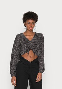 Blusa preta curta com mangas longas, apresentando um padrão de bolinhas brancas dispersas e um decote em V. Combinada com calças pretas de cintura alta.