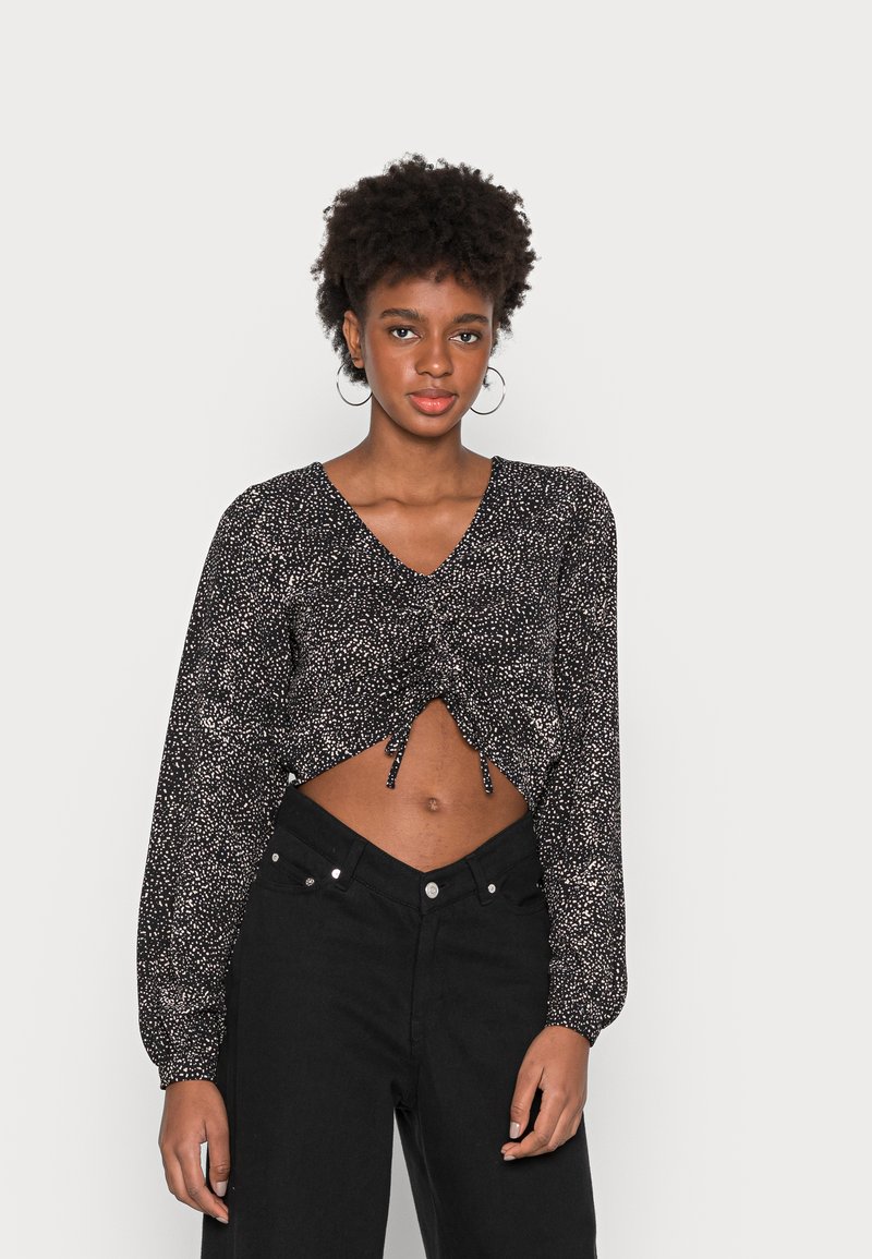 Blusa preta curta com mangas longas, apresentando um padrão de bolinhas brancas dispersas e um decote em V. Combinada com calças pretas de cintura alta.