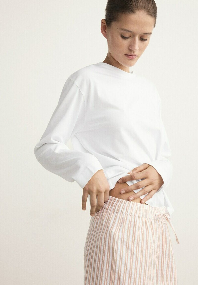 Femme ajustant un pantalon à taille élastique rayé rose pâle et blanc, portant un chemisier blanc uni à manches longues, regardant vers le bas à sa taille.