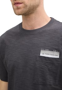 Camiseta de manga corta en gris oscuro, fabricada con material suave y con un cuello redondo; presenta un bolsillo con una etiqueta de marca en el pecho.