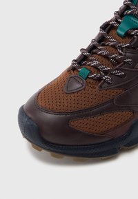 Barna sportcipő lyuggatott hálós és bőr felsőrésszel, türkizkék részlettel, texturált fekete talppal, csúszásgátló mintával és visszaverő fűzőkkel.