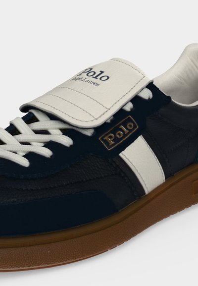 Σκούρο μπλε και λευκό αθλητικό παπούτσι Polo Ralph Lauren με λευκά κορδόνια, καφέ σόλα και ορατή επωνυμία Polo στη γλώσσα και στην πλευρά.
