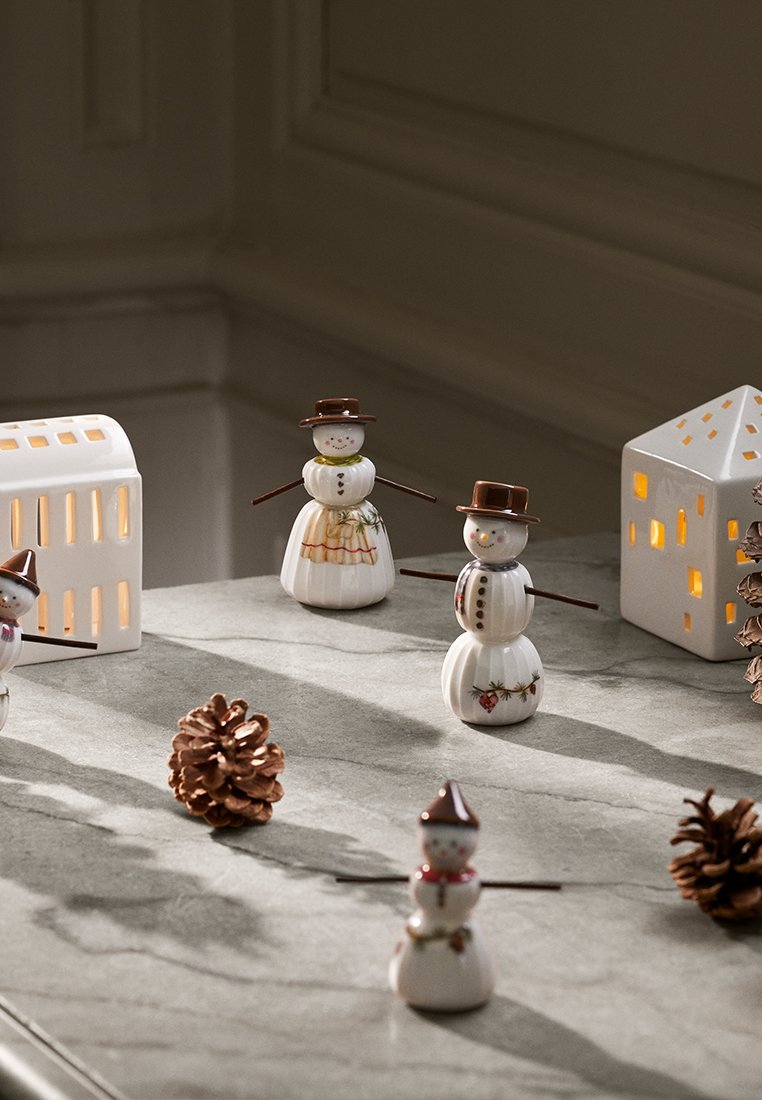 Keramische Schneemänner mit braunen Hüten und Zweigarmen, umgeben von Tannenzapfen und einer weißen laternenförmigen Haus, die auf einer Marmoroberfläche ruhen.