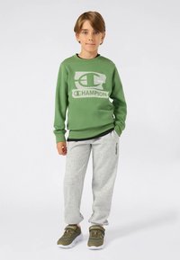 Ragazzo in piedi di fronte, indossa una felpa Champion verde, pantaloni sportivi grigio chiaro Champion e sneakers verdi oliva Champion su sfondo bianco.