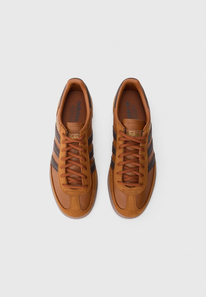 Brown leather sneakers featuring three black stripes, brown laces, and a textured finish. Gold logo accents on the sides and tongue.