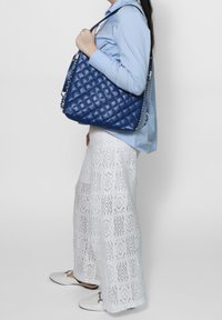 Borsa a tracolla in pelle blu trapuntata con catena, caratterizzata da un design morbido. Indossata sopra una gonna in pizzo bianco e una blusa azzurro chiaro.