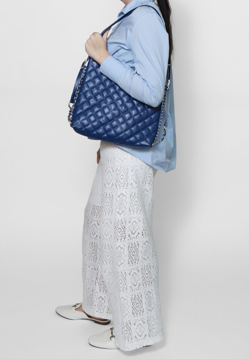 Borsa a tracolla in pelle blu trapuntata con catena, caratterizzata da un design morbido. Indossata sopra una gonna in pizzo bianco e una blusa azzurro chiaro.