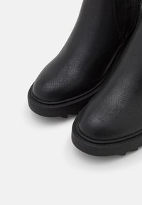 Bottes en cuir noir texturé avec bouts arrondis, semelles épaisses et coutures visibles, présentées en gros plan de face sur un fond blanc.