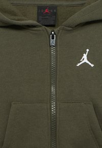 Φούτερ με φερμουάρ Jordan σε ελιά χρώμα, με λευκό λογότυπο Jumpman στο στήθος  και μαύρη ετικέτα που δείχνει το μέγεθος 12M και «Κατασκευάστηκε στο Βιετνάμ».