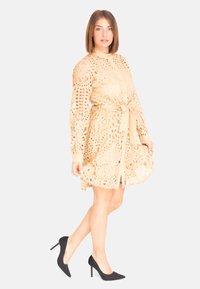 Beige kanten jurk met lange mouwen, een strik taille en ingewikkelde uitsparingen. Gecombineerd met zwarte puntige stilettohakken voor contrast.