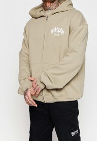 Sudadera beige con cremallera, capucha grande, bolsillos delanteros y puños acanalados. Presenta un logo impreso en blanco en el pecho. Combinada con pantalones cargo negros.