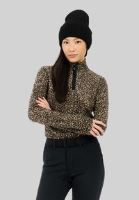 Haut zippé en imprimé léopard en tissu doux, avec un col haut et des manches longues, associé à un pantalon noir ajusté et un bonnet en tricot noir.