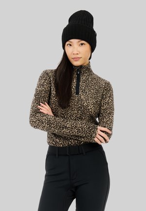Zipperová blúzka s leopardím vzorom z mäkkého materiálu, s vysokým golierom a dlhými rukávmi, kombinovaná s čiernymi priliehavými nohavicami a čiernou pletenou čiapkou.