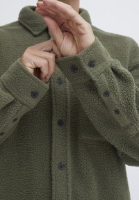 Olivegrønn fleece-skjorte med knappefront, svarte knapper, krage og strukturert stoff. Mansjettene er rullet opp med gravert knappelukking.