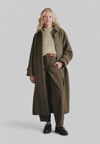 Oliwkowy zielony wełniany płaszcz z kołnierzem i guzikami, w zestawieniu z jasnorozowym zielonym dzianinowym swetrem, brązowymi spodniami ze sztruksu i ciemnymi butami.