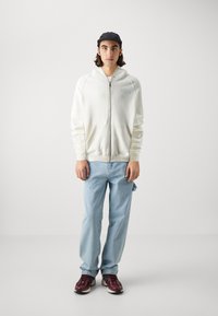 Felpa bianca con zip e logo azzurro chiaro, abbinata a jeans azzurri chiari e sneakers bordeaux. Il modello è in piedi contro uno sfondo semplice.