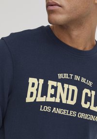Tums zils kokvilnas sporta krekls ar tekstu "BLEND CL" krēmkrāsā, virs kura ir uzraksts "BUILT IN BLUE" un zem kura "LOS ANGELES ORIGINALS".