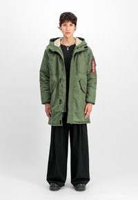 Giacca isolata verde con cappuccio, dotata di fodera color crema, tasche con zip e un'etichetta rossa. Abbinata a pantaloni neri larghi.