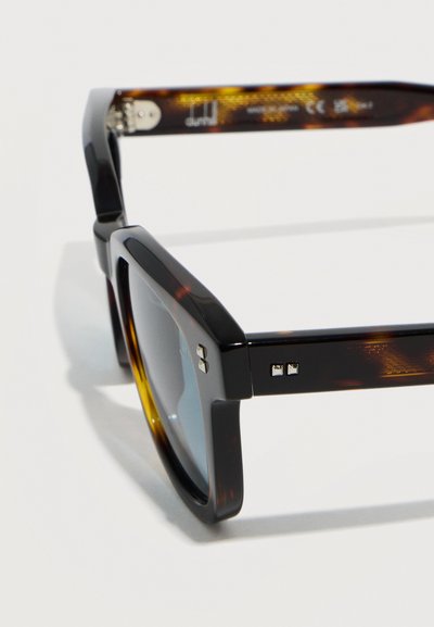 Ορθογώνια γυαλιά ηλίου με σκελετό από χελώνα, παχιές κορνίζες και σκούρους φακούς, εμφανιζόμενα από κοντινή πλευρική γωνία.