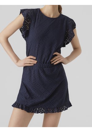 Femme portant une robe bleu marine à œillets, manches courtes volantées et ourlet froncé, mains sur les hanches devant un fond uni.