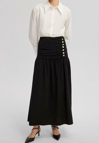 Schwarzer Maxi-Rock mit elastischem Bund und großen goldenen Knöpfen. Kombiniert mit einem weißen Langarmshirt und spitzen schwarzen High Heels.