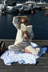 Svetlobéžové pohodlné oblečenie s kvetinovou výšivkou, umiestnené na bielej osuške, s okuliarmi a knihou. Drevené mólo a lode v pozadí.