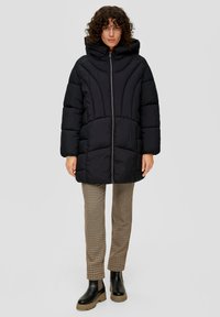 Zwarte gewatteerde jas met capuchon, voorzien van een ritssluiting aan de voorkant, een quiltpatroon en een iets oversized pasvorm. Gecombineerd met geruiten broek en stevige laarzen.