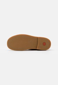 Soletta di scarpa in gomma marrone con superficie testurizzata e accento circolare rosso, marchiata "Kickers". Presenta un design ampio e piatto per una maggiore stabilità.