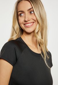 Zwarte top met korte mouwen en ronde hals, gemaakt van een gladde, rekbare stof. Heeft raglanmouwen en een getailleerde pasvorm.