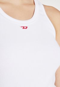 Top alb cu dungi, cu un mic logo brodat roșu aproape de guler. Țesătura pare netedă, având un design ajustat.