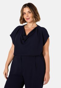 Top blu navy con scollo drappeggiato e maniche corte. Il tessuto è liscio, con una vestibilità rilassata. Abbinato a pantaloni coordinati.