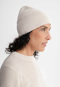 Beige ribgebreide beanie met een ronde bovenkant, gedragen door een persoon met krullend haar, gecombineerd met een gestructureerde beige trui.