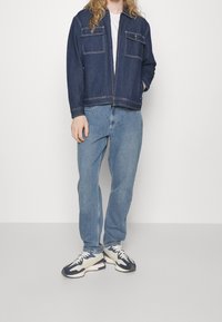 Giacca di jeans con cuciture chiare, due tasche frontali, indossata sopra una maglietta bianca. Abbinata a jeans blu larghi e sneakers multicolori.