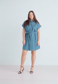 Lys blå denimskjortekjole med korte ermer, knappelukking foran og belte i midjen. Svart høyhælte sandaler fullfører antrekket.