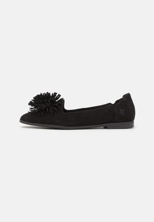Chaussure plate noire avec un grand pompon à l'avant et un logo brodé discret près du talon, présentant un design simple et élégant.