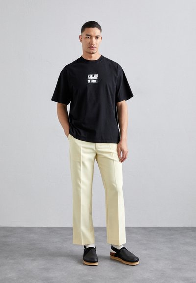 Sort bomulds t-shirt med hvid tekst, gule skræddersyede bukser og sorte slip-on sko med en træsål. Enkel, afslappet pasform.