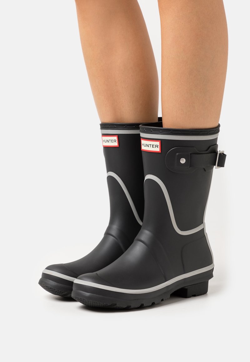 Jambes portant des bottes de pluie en caoutchouc noir mi-mollet avec un bord gris et une boucle latérale, étiquetées Hunter à l’avant.