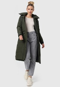 Lange olijfgroene gewatteerde jas met een hoge kraag, ritssluiting aan de voorkant, zijzakken en een gladde textuur, gecombineerd met grijze high-waisted jeans en sneakers.