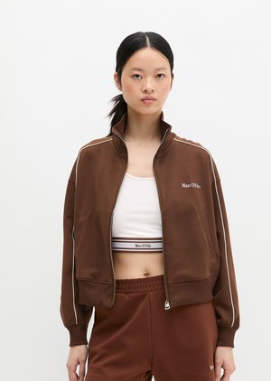 Fiatal nő, fekete hajjal lófarokban, barna cipzáras dzsekit, hozzáillő rövidnadrágot és fehér sportmelltartót, márkajelzésekkel visel.