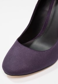 Chaussure à talon haut en daim violet avec une texture lisse, bout pointu et intérieur noir. Élévation et design élégant caractéristiques des escarpins.