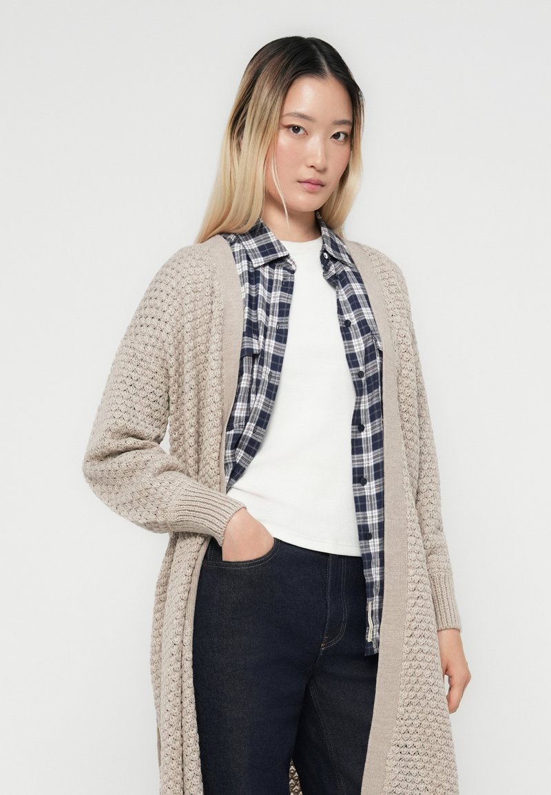 Un cardigan en tricot beige avec un motif texturé, superposé à une chemise à carreaux bleu marine, porté avec un haut blanc et un jean en denim foncé.