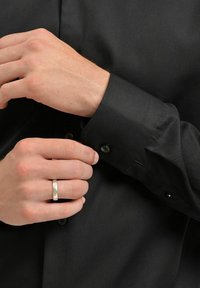 Svart skjorta med knäppning och slätt tyg, med vikta manschetter och svarta knappar. Hand som justerar manschetten, silverring synlig.