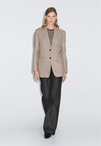 Vrouw in een beige blazer over een zwart-wit gestreept top, gecombineerd met losse zwarte broeken en zwarte schoenen, staande tegen een witte muur.
