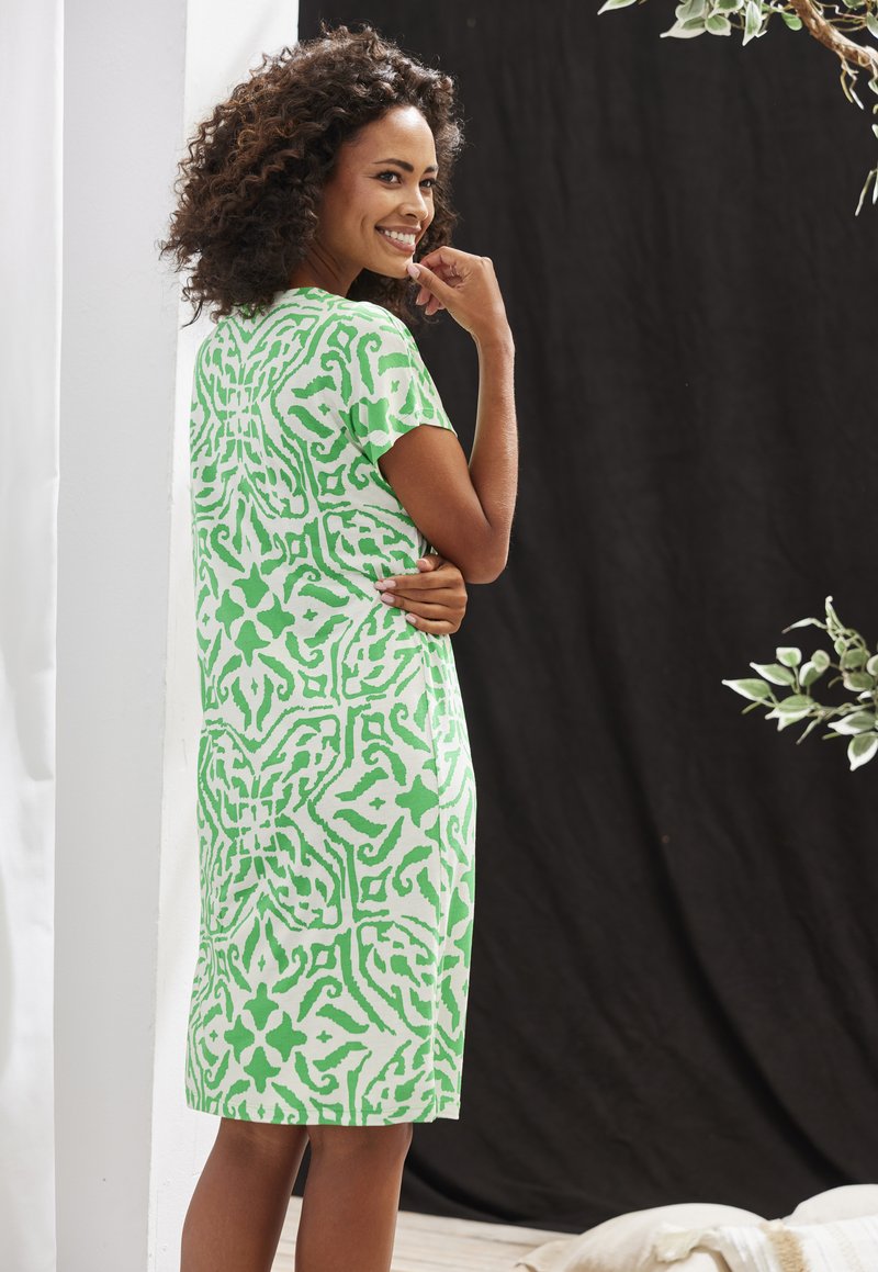 Lyhyt vihreä kuvioitu mekko, jossa on lyhyet hihat ja suora leikkaus, esittää lehväkuviota valkoisella taustalla.