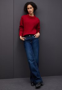 Rode, gestructureerde gebreide trui met een hoge kraag, gecombineerd met donkerblauwe wijde jeans. Zwarte laarzen maken de outfit compleet. Tegen een grijze achtergrond.