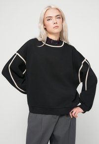 Μαύρη φούτερ μπλούζα με κρεμ λεπτομέρειες, σε χαλαρή γραμμή, με φαρδιές μανσέτες και ριμπ λαιμόκοψη. Συνδυάζεται με γκρι παντελόνι.