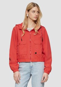 Rote kurz geschnittene Jacke mit Kapuze, Frontreißverschluss, elastischen Bündchen und Saum, mit zwei Brusttaschen. Trägt man über einem schwarz-weiß gestreiften Shirt.