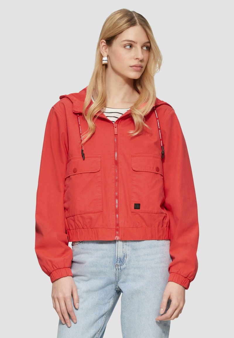 Rote kurz geschnittene Jacke mit Kapuze, Frontreißverschluss, elastischen Bündchen und Saum, mit zwei Brusttaschen. Trägt man über einem schwarz-weiß gestreiften Shirt.