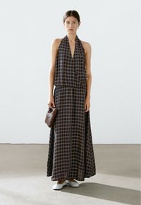 Maxi dress with a halter neckline, featuring a blue and brown checkered pattern. Lightweight fabric, cinched waist, and side slits. White flats and a woven brown bag.